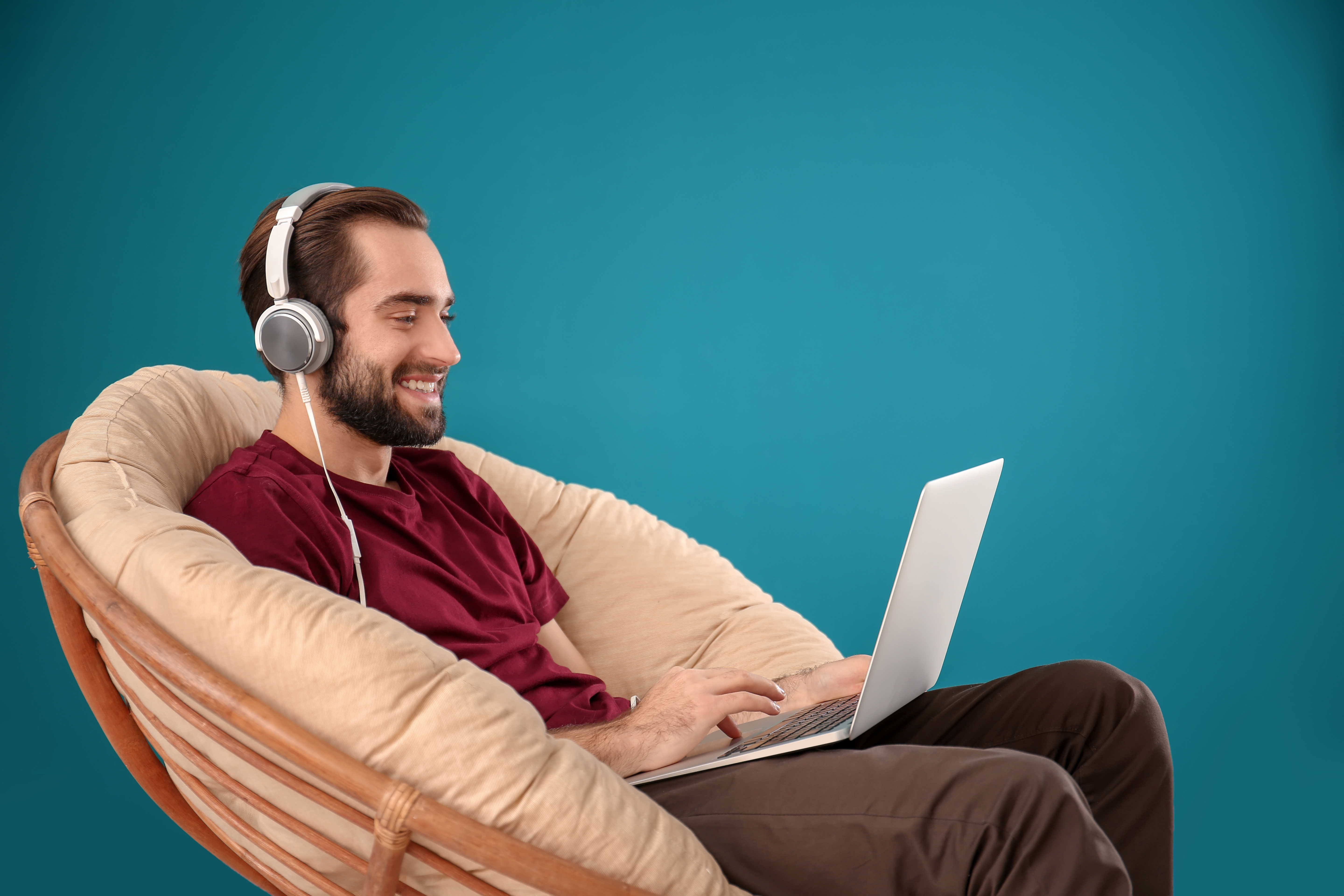 handsome-man-with-laptop-listening-to-music-while-sitting-in-comfortable-armchair-against-color-wall